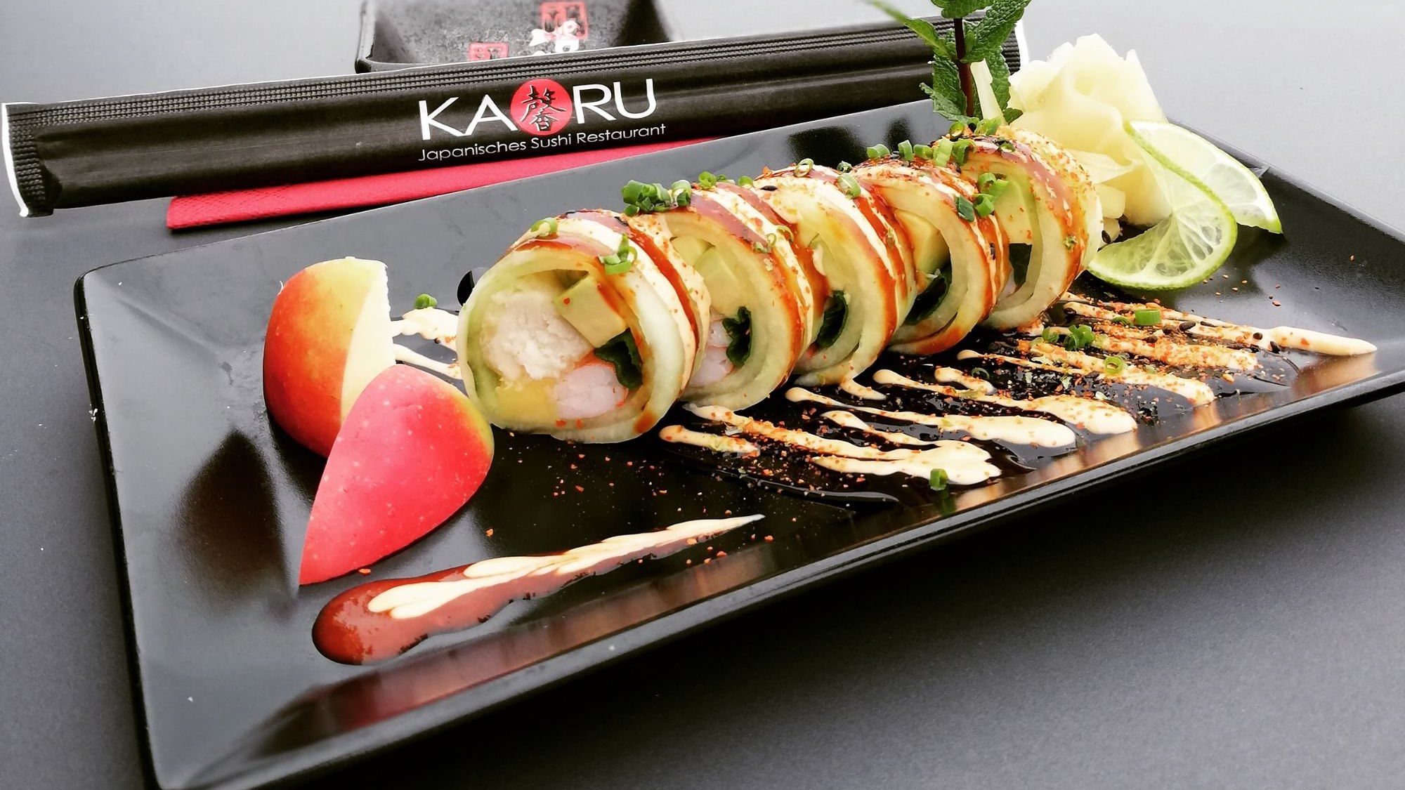 Sushi-Rolle im Gurkenmantel mit Apfel, Limette und Saucen auf schwarzer Platte, daneben KAORU Stäbchen